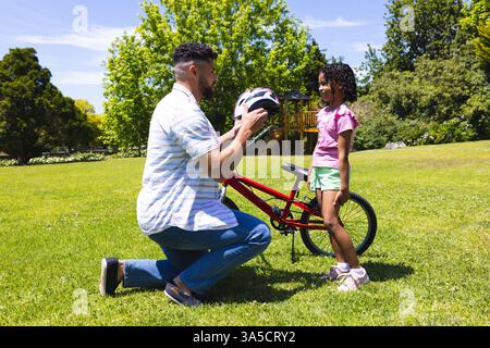 Père aidant la fille avec casque avant de faire du vélo dans le parc ensoleillé Banque D'Images