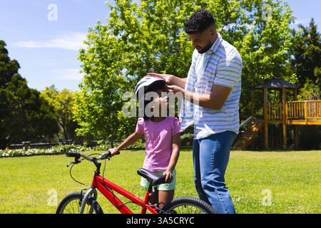Père aidant la fille avec casque avant de faire du vélo dans le parc ensoleillé Banque D'Images