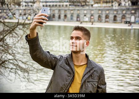 Trois quarts de longueur de jeune homme brun clair aux cheveux portant une veste grise et un Jean denim prenant une photo selfie avec son smartphone devant imageq Banque D'Images