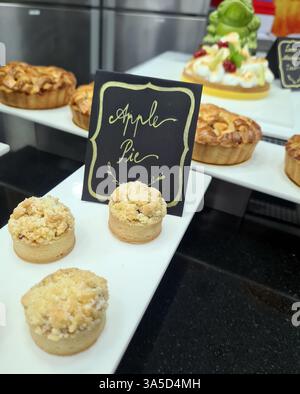 Une tarte aux pommes fraîchement cuite et des mini-pâtisseries sont habilement disposées sur un présentoir blanc dans une boulangerie locale, invitant les clients à se faire plaisir. Banque D'Images