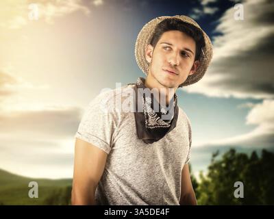 Beau jeune homme aux yeux verts avec chapeau de paille et bandana noir, dehors au coucher du soleil Banque D'Images