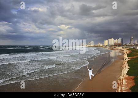 Nuage d'orage suspendu au-dessus de la mer, la femme en blanc exécute Asana Tree sur une jambe. Promenade et plage à tel Aviv Banque D'Images