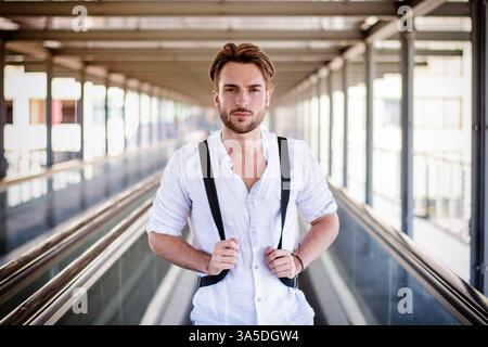 Beau jeune homme marchant en ville avec sac à dos sur les épaules, portant une chemise blanche Banque D'Images