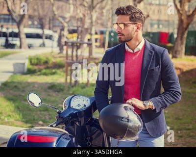 Jeune homme élégant en costume et lunettes de soleil debout par son scooter italien classique sur la route vide dans la ville ensoleillée Banque D'Images
