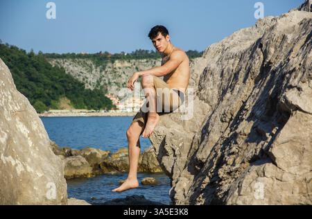 Attrayant beau jeune homme assis sur un rocher par mer ou ocean shore avec les cheveux mouillés, regardant au loin, vue de profil Banque D'Images