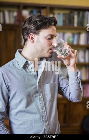 Jeune homme en chemise buvant de l'eau de verre de cristal sur fond d'étagères Banque D'Images