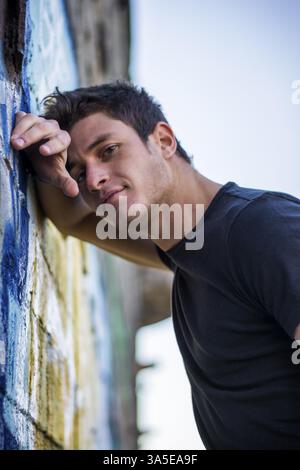 Séduisant jeune homme debout appuyé contre un mur de graffitis colorés, looking at camera Banque D'Images