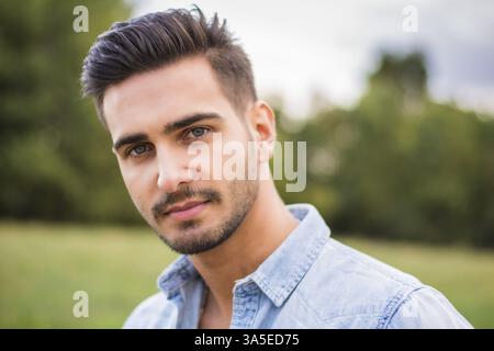 Beau jeune homme à la campagne, devant un champ ou une prairie, portant une chemise, regardant loin d'un côté Banque D'Images
