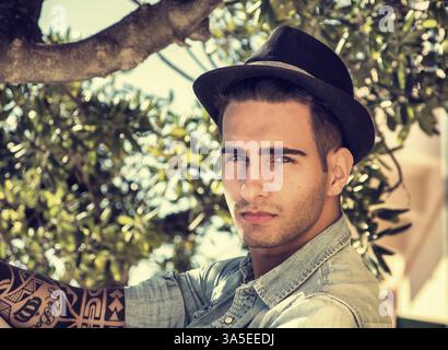 Beau jeune homme contre les branches d'arbres d'olive, looking at camera, dans une journée ensoleillée, le port de fedora hat Banque D'Images