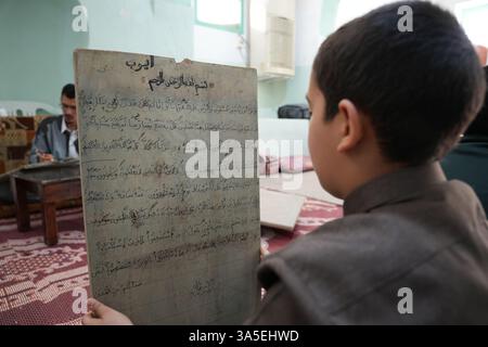 Misrata, Libye. 23 mars 2025. Un étudiant se concentre sur la lecture de versets du Saint Coran écrits sur sa tablette en bois, les enfants libyens sont assis dans une école coranique traditionnelle, inscrivant soigneusement des versets sur des tablettes en bois pendant que leur professeur les guide dans la récitation. Cette méthode séculaire, ravivée pendant le Ramadan, reste une pratique chère pour mémoriser le Saint Coran. (Photo par Islam Alatrash/SOPA images/SIPA USA) crédit : Sipa USA/Alamy Live News Banque D'Images