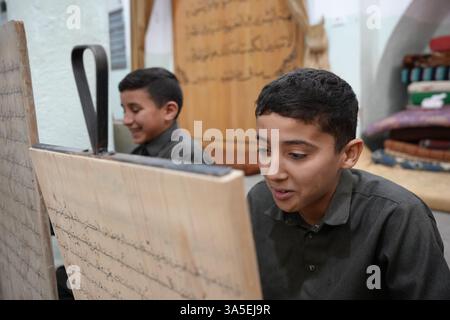 Misrata, Libye. 23 mars 2025. Un étudiant se concentre sur la lecture de versets du Saint Coran écrits sur sa tablette en bois, les enfants libyens sont assis dans une école coranique traditionnelle, inscrivant soigneusement des versets sur des tablettes en bois pendant que leur professeur les guide dans la récitation. Cette méthode séculaire, ravivée pendant le Ramadan, reste une pratique chère pour mémoriser le Saint Coran. (Photo par Islam Alatrash/SOPA images/SIPA USA) crédit : Sipa USA/Alamy Live News Banque D'Images