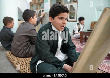 Misrata, Libye. 23 mars 2025. Un étudiant se concentre sur la lecture de versets du Saint Coran écrits sur sa tablette en bois, les enfants libyens sont assis dans une école coranique traditionnelle, inscrivant soigneusement des versets sur des tablettes en bois pendant que leur professeur les guide dans la récitation. Cette méthode séculaire, ravivée pendant le Ramadan, reste une pratique chère pour mémoriser le Saint Coran. Crédit : SOPA images Limited/Alamy Live News Banque D'Images