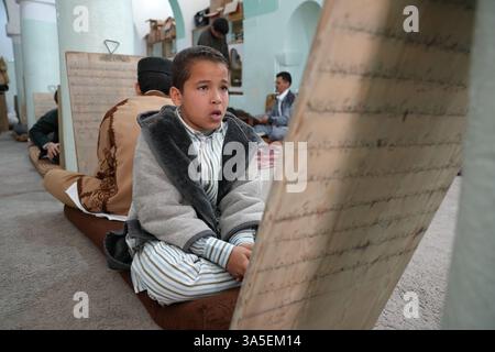 Misrata, Libye. 23 mars 2025. Un étudiant se concentre sur la lecture de versets du Saint Coran écrits sur sa tablette en bois, les enfants libyens sont assis dans une école coranique traditionnelle, inscrivant soigneusement des versets sur des tablettes en bois pendant que leur professeur les guide dans la récitation. Cette méthode séculaire, ravivée pendant le Ramadan, reste une pratique chère pour mémoriser le Saint Coran. Crédit : SOPA images Limited/Alamy Live News Banque D'Images