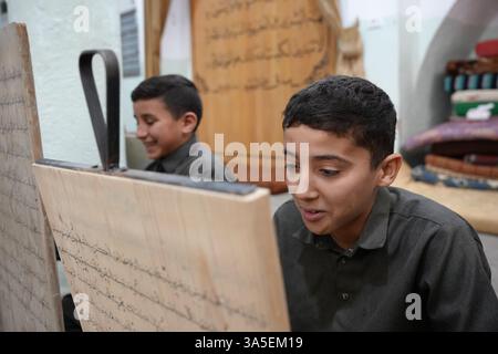 Misrata, Libye. 23 mars 2025. Un étudiant se concentre sur la lecture de versets du Saint Coran écrits sur sa tablette en bois, les enfants libyens sont assis dans une école coranique traditionnelle, inscrivant soigneusement des versets sur des tablettes en bois pendant que leur professeur les guide dans la récitation. Cette méthode séculaire, ravivée pendant le Ramadan, reste une pratique chère pour mémoriser le Saint Coran. Crédit : SOPA images Limited/Alamy Live News Banque D'Images