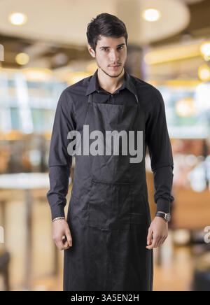 Jeune cuisinier ou serveur posant, le port de tablier noir et chemise en bar ou restaurant ou dîner Banque D'Images