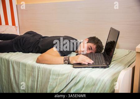 Close Up of assez jeune homme aux cheveux noirs de dormir sur le plancher à côté d'un ordinateur portable avec la main sur le clavier Banque D'Images