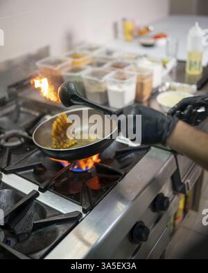 Cuisinier professionnel portant des gants noirs, ajoutant des pois chiches dans une poêle remplie d'épices aromatiques, préparant habilement la cuisine indienne traditionnelle dans un Banque D'Images