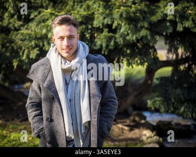 Beau jeune homme à la mode, se tenir debout ou marcher dans le parc de la ville à la mode de porter un chandail ou un cardigan et foulard Banque D'Images