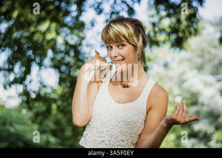 Jolie blonde woman m'appeler signe avec la main, comme un symbole de la communication, de l'outdoor dans la nature, avec des arbres derrière Banque D'Images