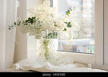 Bouquet de belles fleurs de gypsophila et branches d'eucalyptus dans un vase sur le rebord de la fenêtre à l'intérieur Banque D'Images