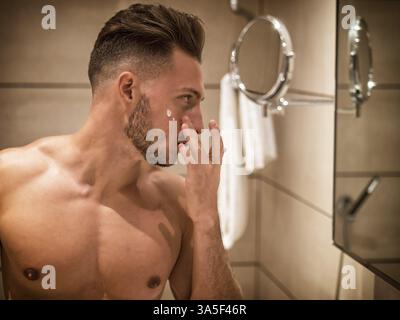 Beau jeune homme appliquant une crème hydratante sur la peau de son visage, à la maison dans sa salle de bain Banque D'Images