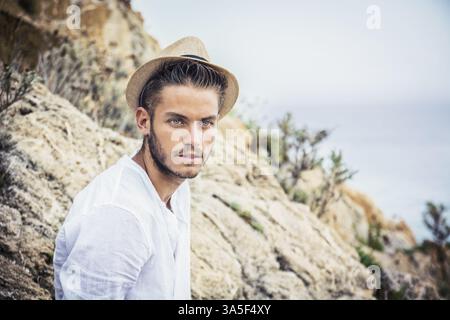 Beau jeune homme dans le quartier branché d'accoutrement, sur une plage dans une journée ensoleillée, portant une chemise blanche et chapeau de paille Banque D'Images