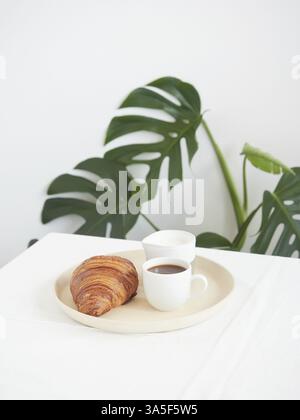 Petit déjeuner encore en vie. Croissant, tasse à café et pot à lait sur la table, plante à feuilles vertes en arrière-plan Banque D'Images