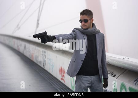 Bien habillé beau jeune détective ou policier ou gangster debout dans un environnement urbain visant une arme au côté gauche Banque D'Images