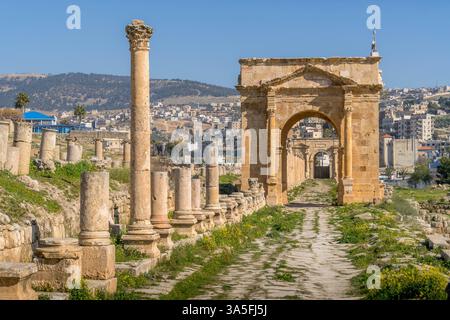 Ruines romaines historiques à Jerash, en Jordanie, présentant une rue à colonnades et une grande arche, situées contre des collines ondulantes et un paysage urbain moderne. Banque D'Images