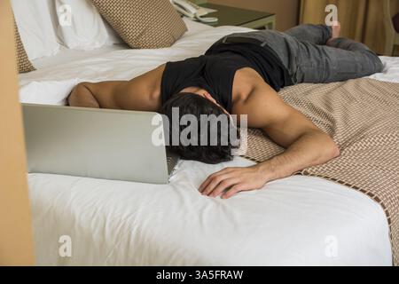 Close Up of assez jeune homme aux cheveux noirs de dormir sur le plancher à côté d'un ordinateur portable avec la main sur le clavier Banque D'Images