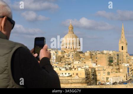 Un touriste âgé aux cheveux gris portant des lunettes de soleil prend des photos avec son téléphone vu d'en haut, capturant une vieille ville balnéaire. Banque D'Images
