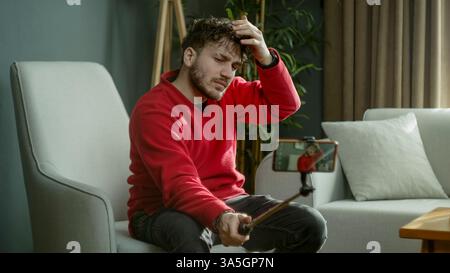 Jeune homme dans un pull rouge utilisant un bâton selfie pour enregistrer une vidéo ou prendre une photo tout en étant assis dans un salon moderne Banque D'Images
