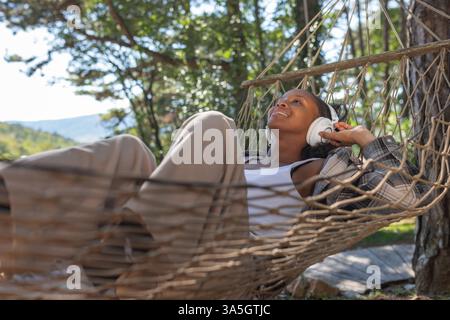 Insouciante jeune femme afro-américaine se relaxant sur un hamac, appréciant écouter de la musique à travers des écouteurs sans fil, et la belle nature autour Banque D'Images