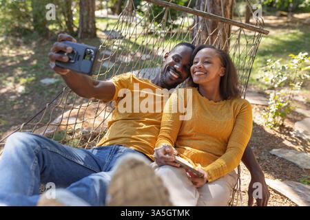 Couple afro-américain souriant insouciant passant leur journée de vacances à se détendre dans un hamac, profitant d'un magnifique environnement naturel de site de glamping. Banque D'Images