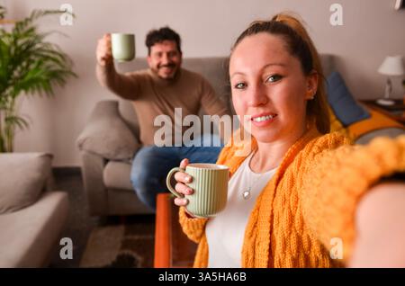Jeune couple prenant un selfie tout en buvant un café sur leur canapé à la maison Banque D'Images