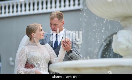 Heureux jeunes mariés se tenant la main et se regardant près d'une fontaine, célébrant leur mariage Banque D'Images
