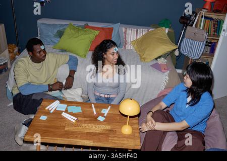 Trois amis appréciant de jouer au jeu dans un salon confortable avec des notes autocollantes sur leurs fronts, indiquant une interaction vive et engageante les uns avec les autres Banque D'Images