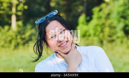 Portrait d'une jeune femme joyeuse avec des lunettes de soleil, profitant d'une journée ensoleillée dans un parc, souriant et tenant son menton Banque D'Images