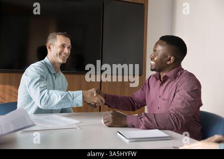 Joyeux partenaires d'affaires masculins multiethniques serrant la main à la table de conférence, souriant, finissant la conversation, la négociation ou consultati professionnel Banque D'Images