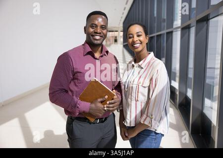 Portrait de bureau de deux jeunes membres du personnel africains debout dans le couloir moderne souriant à la caméra, exsudant compétence, confiance, ayant positif et c Banque D'Images