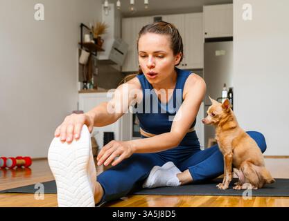 femme assise sur le tapis de yoga exercice d'étirement à la maison tandis que petit chien la regardant Banque D'Images