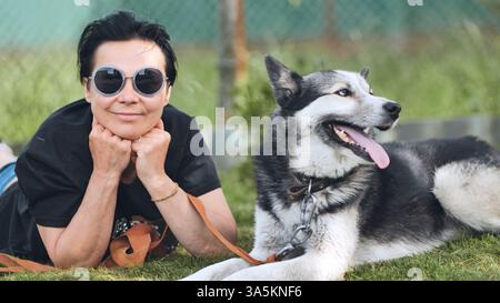 Heureuse femme mature portant des lunettes de soleil couchées sur l'herbe avec son chien Malamute d'alaska, profitant du temps libre dans un jardin Banque D'Images