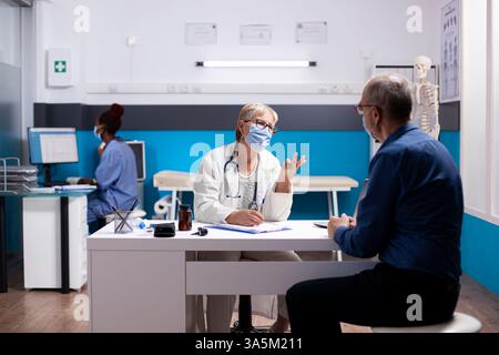 L'homme retraité est assis à son bureau, discutant des détails des soins de santé avec une vieille médecin en blouse de laboratoire, examinant les notes de traitement. Le médecin âgé utilise le presse-papiers pour partager les résultats médicaux avec le patient retraité. Banque D'Images