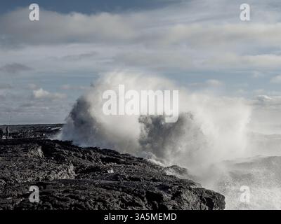 D'énormes vagues s'écrasent contre des roches de lave déchiquetées en Islande comme le montre une figure lointaine. Un moment étonnant de contraste entre présence humaine et natur Banque D'Images