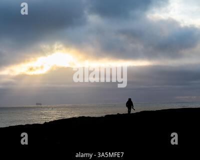 Un voyageur solitaire marche le long de la côte sombre de Valahnúkamöl, en Islande, sous un ciel sombre tandis que les rayons du soleil traversent les nuages et scintillent au-dessus du nord Banque D'Images