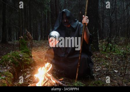 Un moine aux tons noirs est assis près d'un feu dans la forêt et regarde dans une boule de divination. Mysticisme et magie, sorciers et sorcières. Banque D'Images
