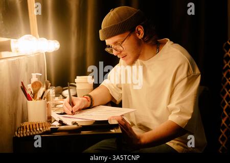 Le jeune homme s'est concentré sur l'étude et l'écriture de notes au bureau sous la lumière chaude de la lampe, portant un bonnet et des lunettes. Entouré de matériel d'étude et d'objets personnels Banque D'Images