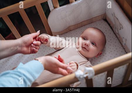 Une mère aimante tient doucement les mains de son bébé allongé dans un berceau, créant une tendresse Banque D'Images
