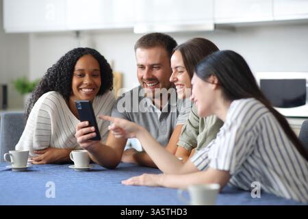 Quatre amis interraciaux heureux parlant du contenu du téléphone dans la cuisine à la maison Banque D'Images
