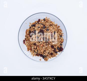 Un bol en verre transparent rempli de granola, avec un mélange d'avoine, de noix et de fruits secs comme des raisins secs et des canneberges, sur fond blanc. Banque D'Images